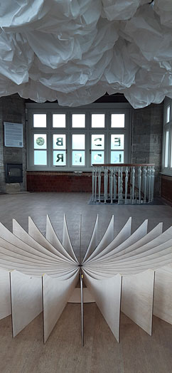 The interior of a high-ceilinged rectangular room with windows on three sides.  In the foreground is a large, circular floor-based sculpture made of plywood. Cloud-like forms hang above it.